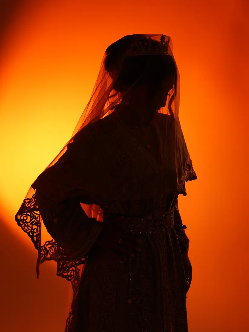 Silhouette voilée en pied, main sur la hanche, en contre-jour sur fond orange.